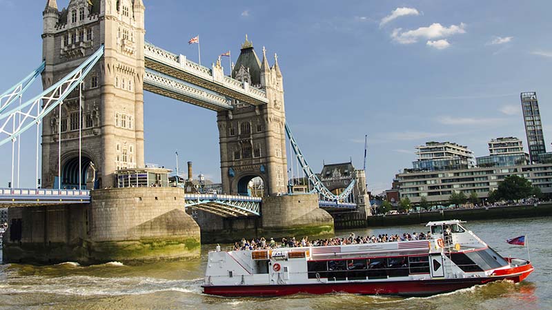 A tour boat travels on the River Thames in front of London's Tower Bridge on a sunny day, with modern buildings in the background and blue sky above.