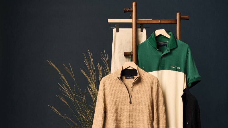 Clothing display against a dark backdrop shows beige pants, a beige textured zipper sweater, and a green and cream polo. Decorative plants add a natural touch.