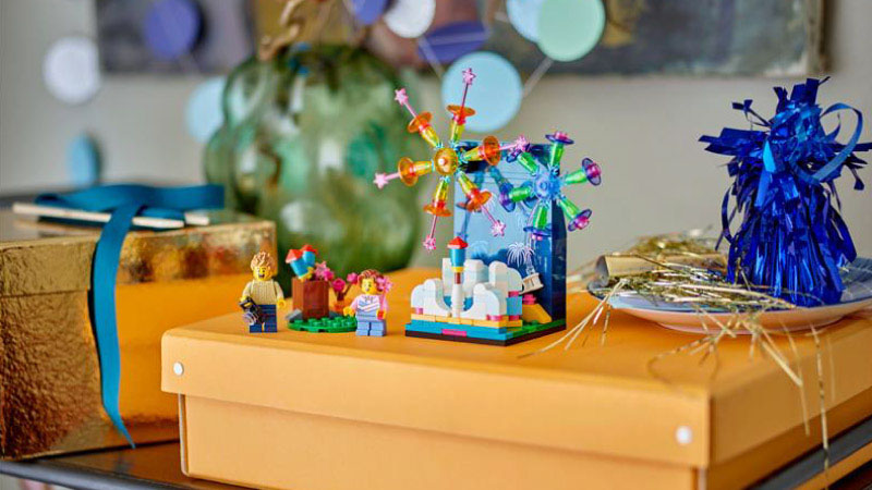 Festive scene with colorful LEGO figures and a Ferris wheel on an orange box. Gift boxes with ribbons and shiny decorations set a celebratory tone.