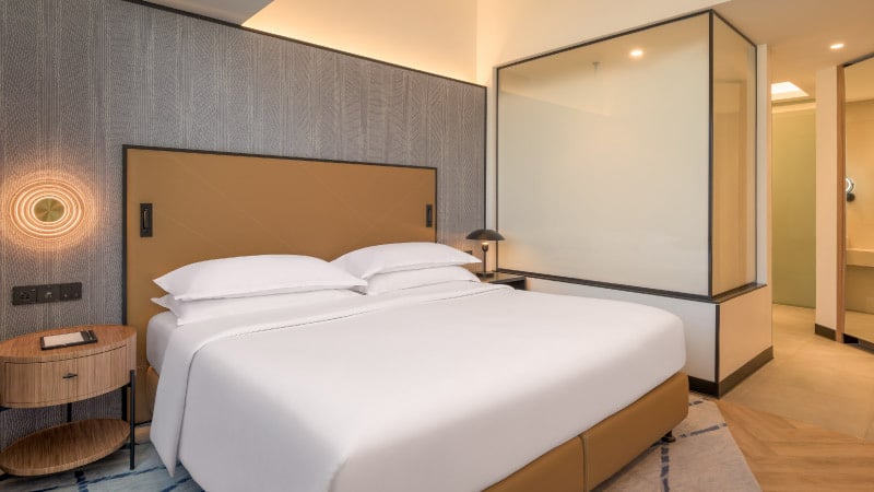 Modern hotel room with a large, neatly made bed with white linens. A wooden nightstand beside it holds a lamp. Glass panel partially divides the room.