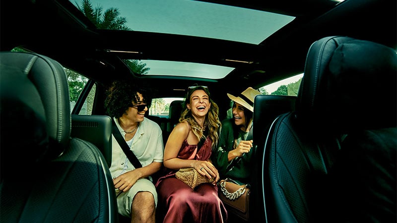 Three people sit in the backseat of a car with a panoramic sunroof, smiling and laughing together.