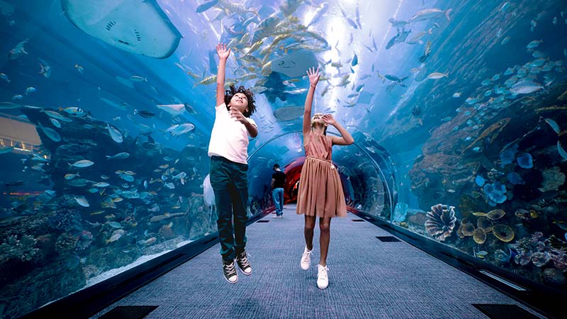 Two children jump with arms raised inside an aquarium tunnel surrounded by fish and marine life, with people visible in the background.