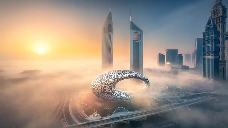 Futuristic silver loop building with Arabic calligraphy amidst fog in a cityscape at sunrise, flanked by modern skyscrapers, conveying calm